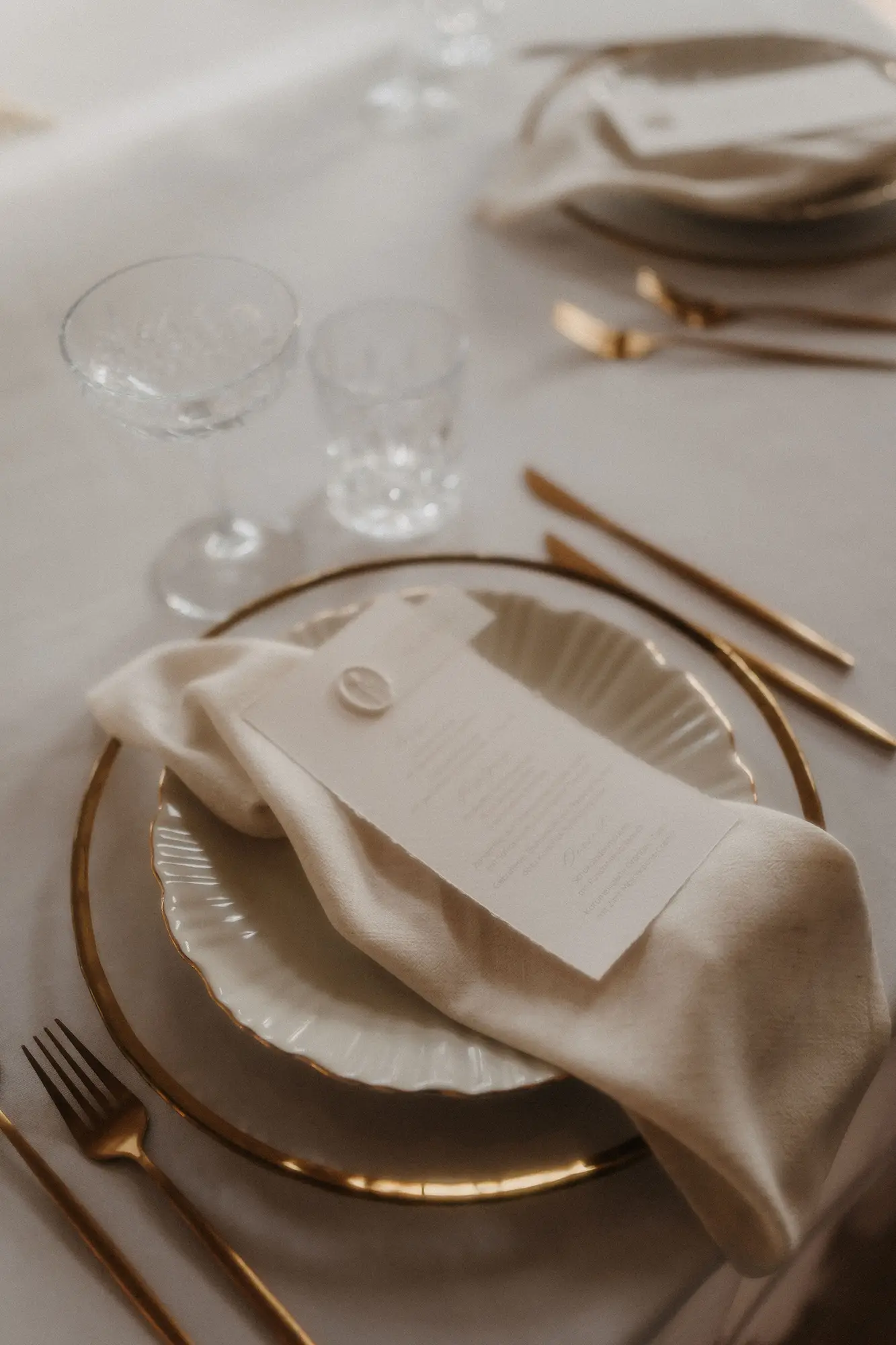 Elegant gedeckter Tisch mit einem weißen Teller mit Goldrand, goldenem Besteck, einer gefalteten elfenbeinfarbenen Serviette, einer bedruckten Menükarte und zwei klaren Gläsern auf einer weißen Tischdecke - gestaltet von Dominique Kubicek für exquisite hochzeitsfloristik in Niederbayern.