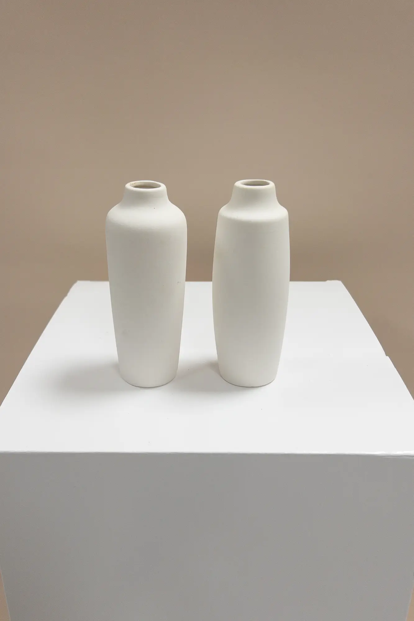 Zwei kleine, weiße, minimalistische Keramikvasen stehen nebeneinander auf einem weißen Sockel vor einem unifarbenen beigen Hintergrund und fangen die Essenz der modernen Hochzeitsfloristik für eine elegante Event & Floral Design Hochzeit Inspiration ein.