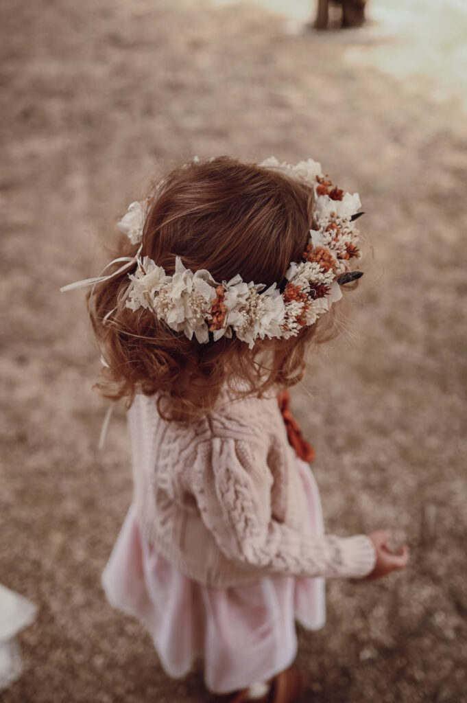 Ein junges Mädchen mit lockigem Haar trägt einen cremefarbenen Pullover und ein rosafarbenes Kleid, das mit einer Blumenkrone im Stil der modernen Hochzeitsfloristik gekrönt ist, und steht im Freien auf einem Kiesweg.