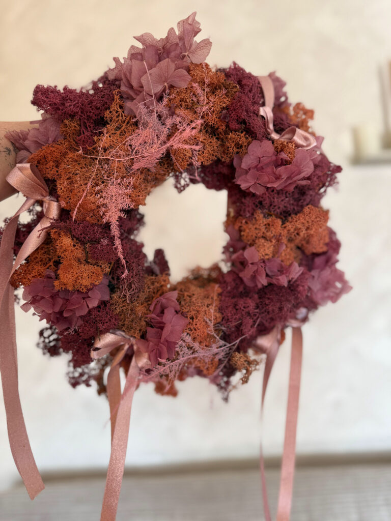 Ein Kranz aus getrockneten, burgunderroten und rosafarbenen Blumen und Moos, verziert mit rosafarbenen Bändern, wird vor einem weichen, neutralen Hintergrund gehalten - ein zartes Stück Hochzeitsfloristik perfekt für Ihr Event & Floral Design Hochzeit by mondhaende.