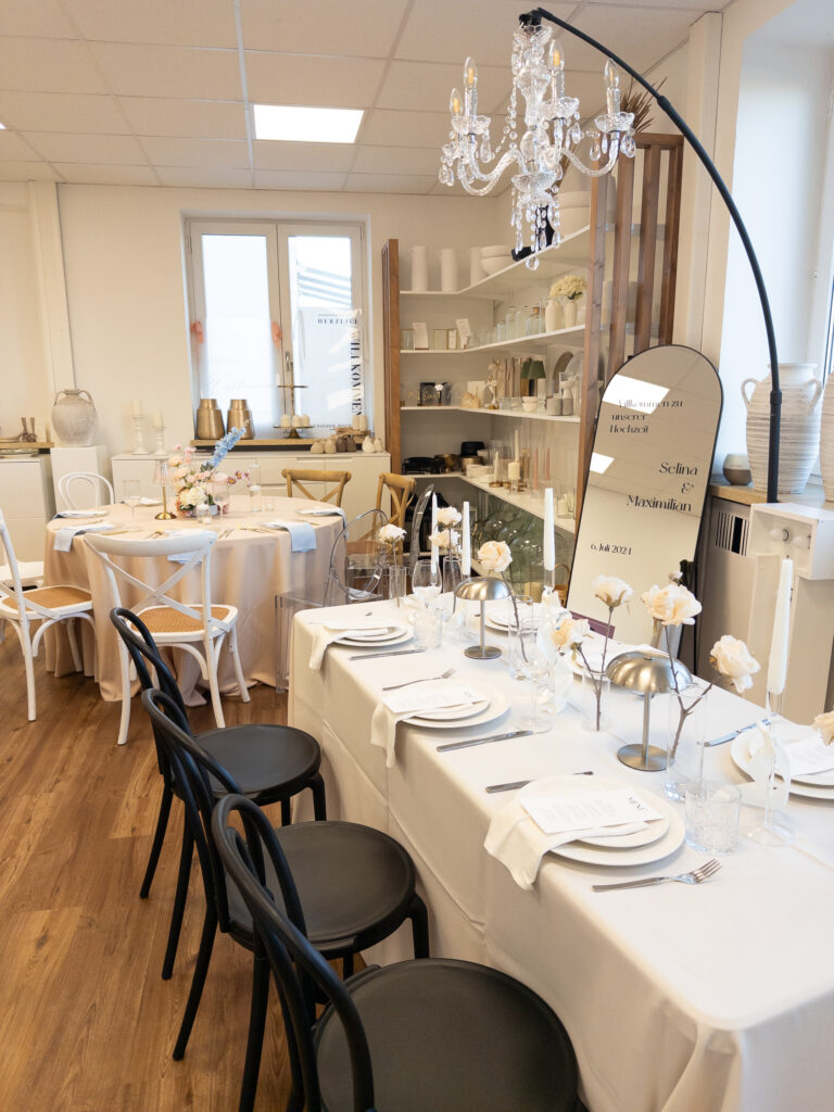 Ein stilvolles Event-Setup mit eleganten weißen Tischen, schwarz-weißen Stühlen, Kerzen, Glaswaren und dekorativen Blumen von hochzeitsfloristik. Darüber hängt ein Kronleuchter, und ein großer geschwungener Spiegel steht neben Keramik in einem hellen Raum.