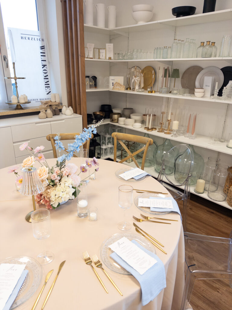 Ein runder Tisch mit beiger Tischdecke ist für vier Personen gedeckt, mit goldenem Besteck, Glastellern, Speisekarten und einem mondänen floralen Mittelstück von Dominique Kubicek Event & Floral Design Hochzeit. Dahinter befinden sich Regale mit Glaswaren, Vasen, Kerzen und Dekoration.