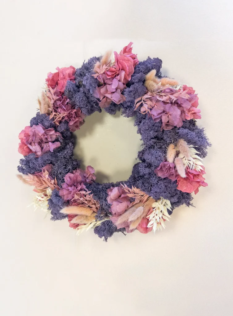 Ein farbenfroher Blumenkranz von Dominique Kubicek mit lilafarbenem Moos, getrockneten rosa und lilafarbenen Blumen und hellbraunen Gräsern präsentiert moderne Hochzeitsfloristik vor einem schlichten hellen Hintergrund. Perfekt für Event & Floral Design Hochzeit.