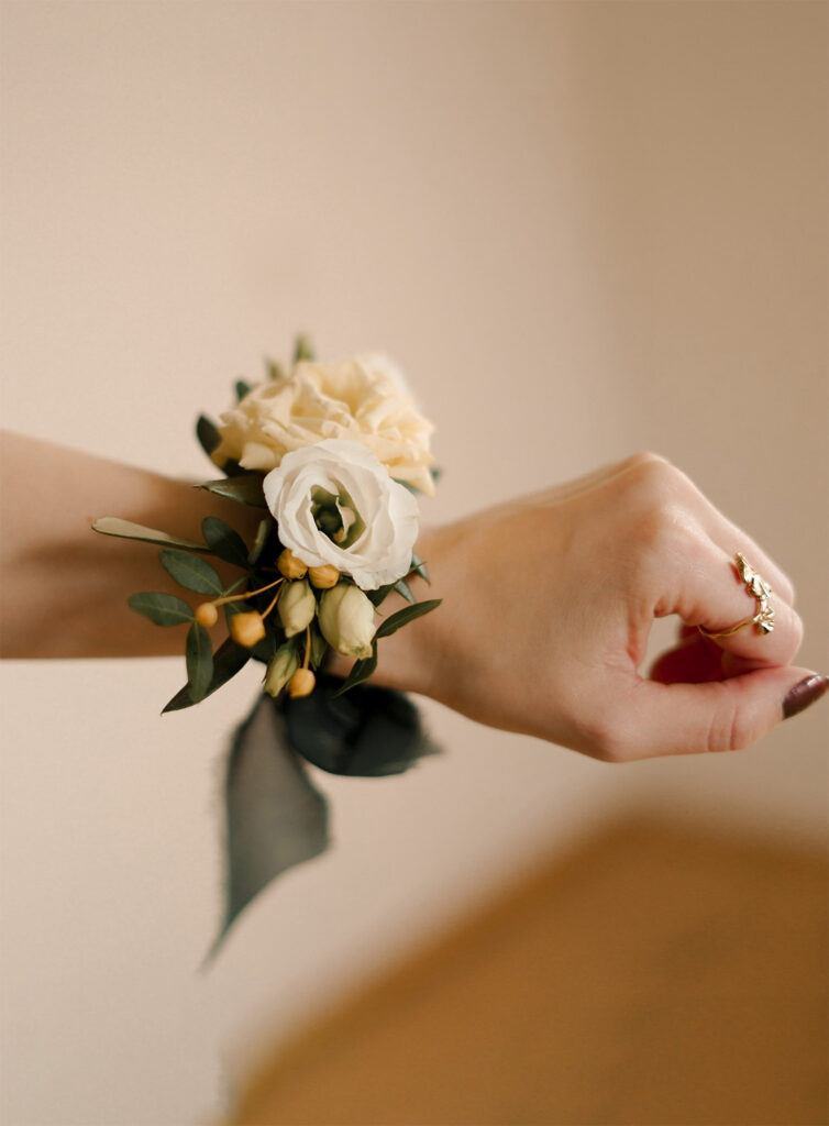 Eine Hand, die einen goldenen Ring und eine zarte florale Handgelenkskorsage mit weißen und hellgelben Blumen trägt, entworfen von Dominique Kubicek Event & Floral Design Hochzeit, vor einem schlichten, sanft verschwommenen Hintergrund in Niederbayern.