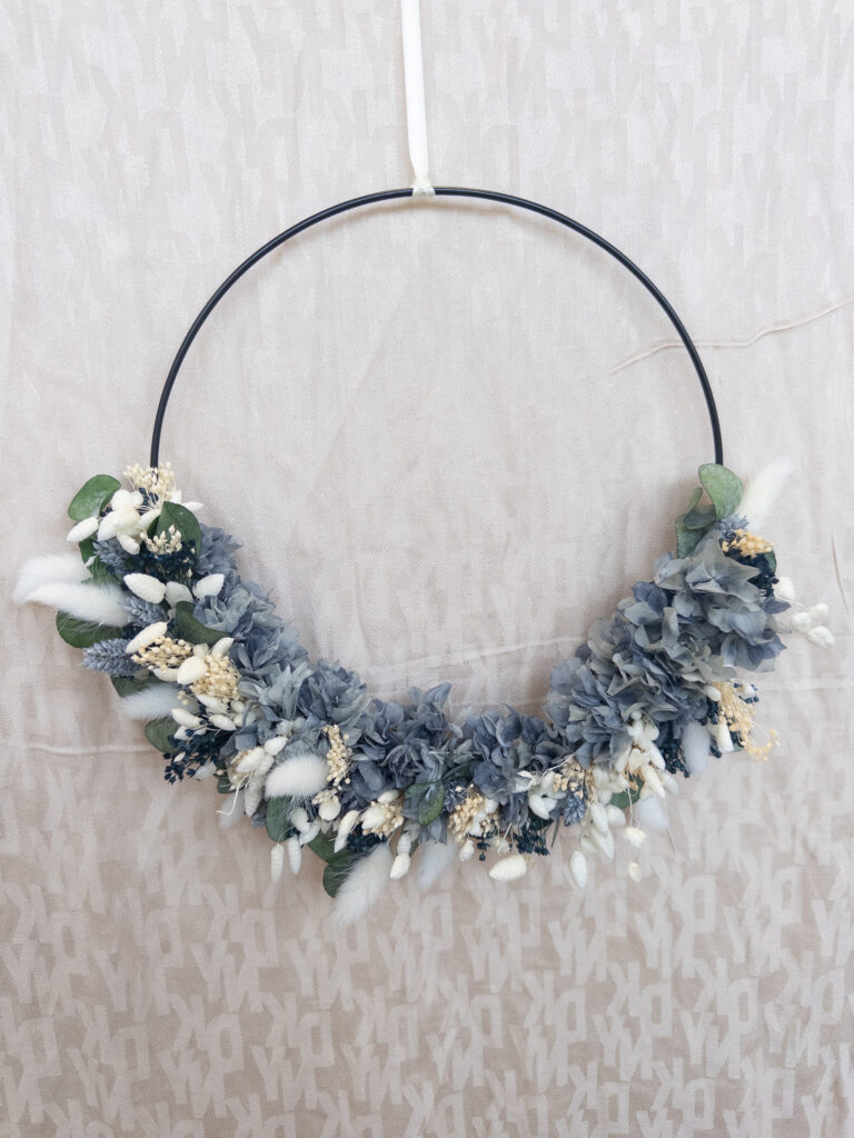 Ein schwarzer Reifenkranz aus Metall, geschmückt mit blauen, weißen und cremefarbenen Kunstblumen und Laub, inspiriert von der modernen Hochzeitsfloristik, auf einem hell gemusterten beigen Stoffhintergrund.